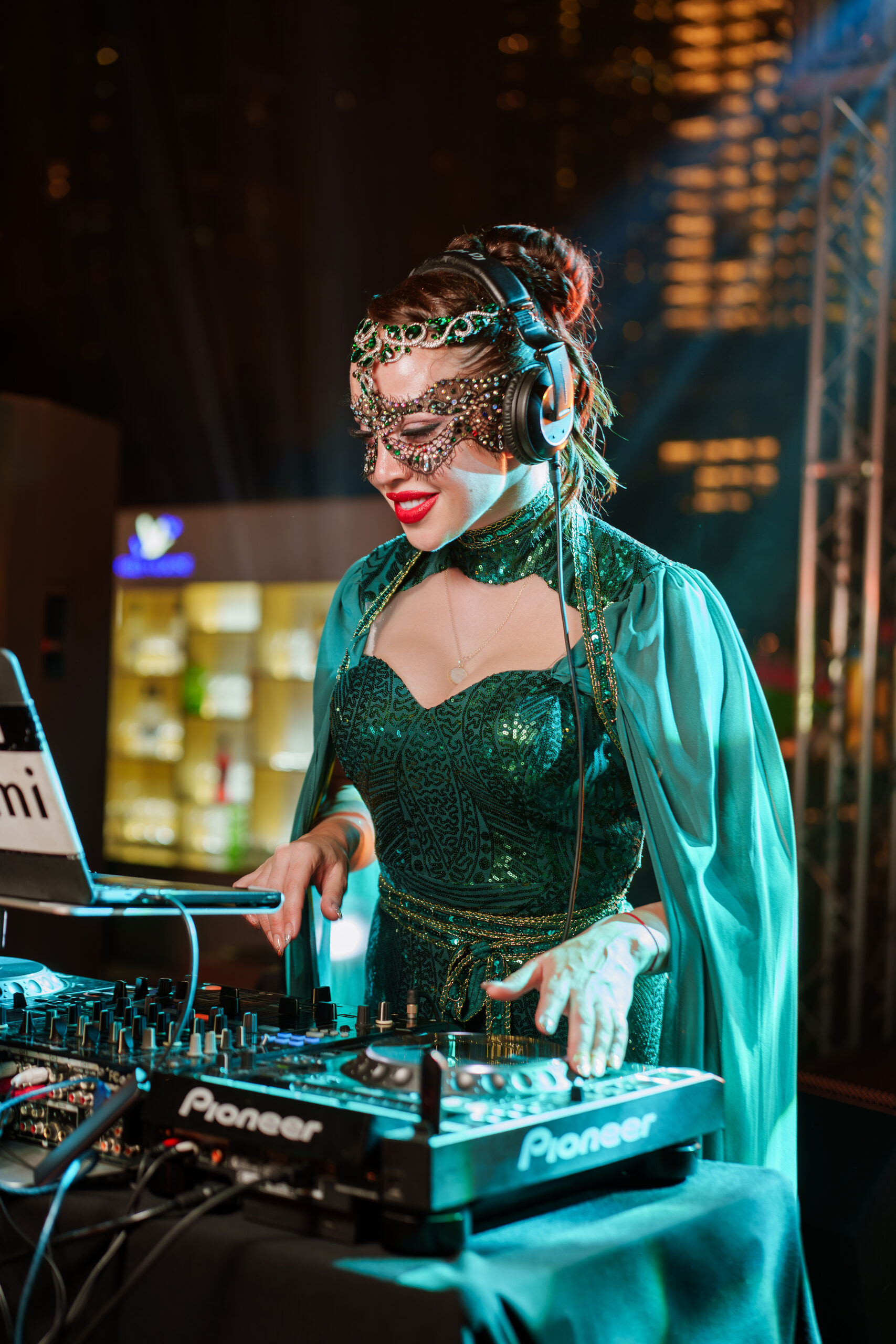 DJ in a jewelled mask and green gown mixing on Pioneer decks at a rooftop event